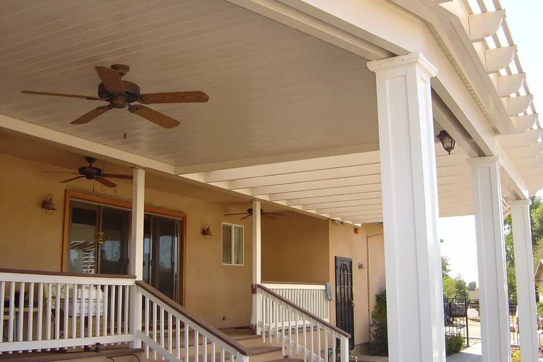 Covered patio with white columns, ceiling fans, tan walls, and white railings beside a house