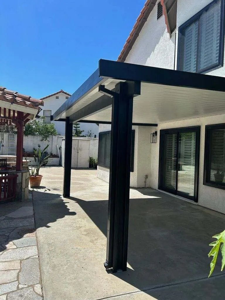 Modern black patio cover with white underside, supported by posts beside a stucco home and concrete patio