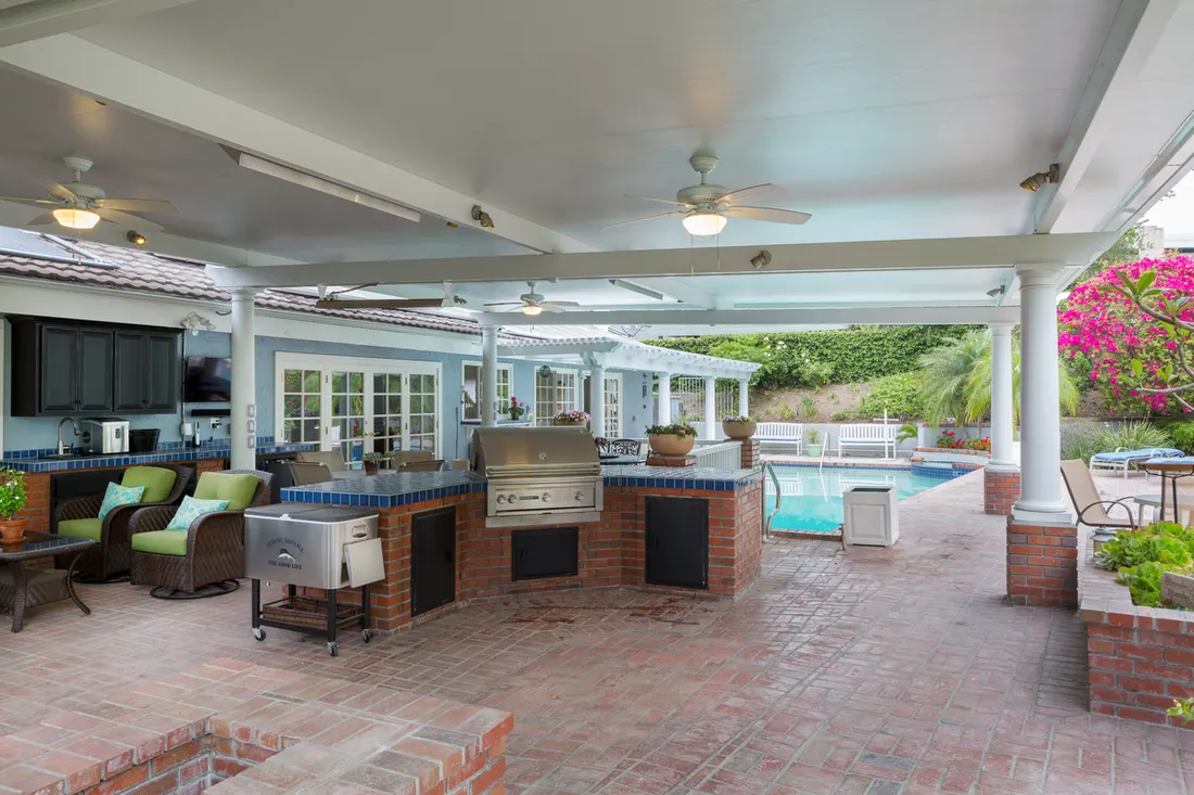 Covered brick patio with white ceiling, outdoor kitchen, pool, and flowering greenery