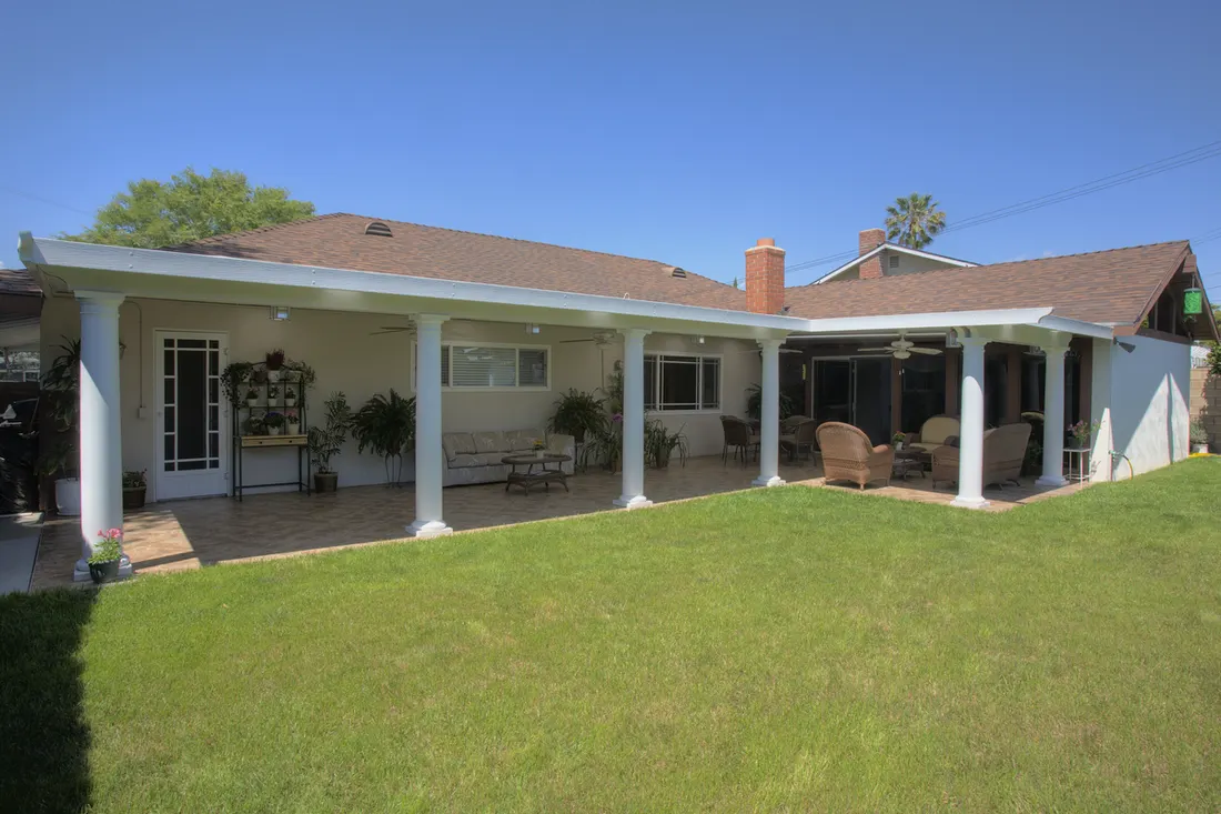 White covered patio with round columns sheltering an outdoor tiled seating area beside a lawn