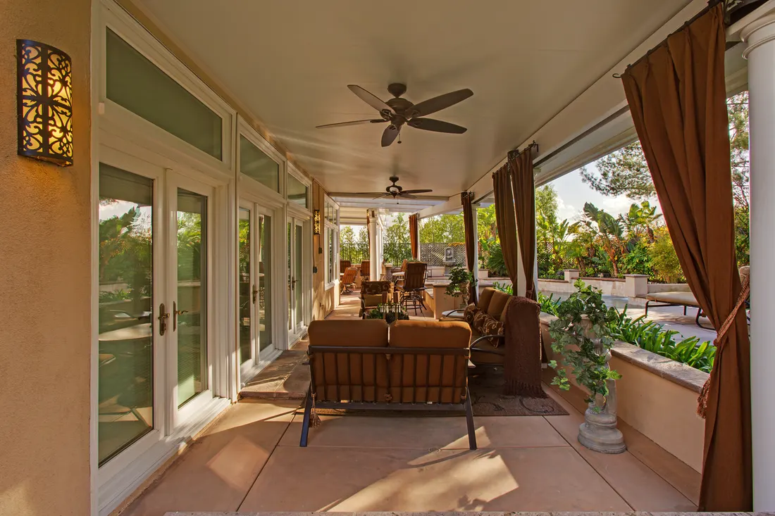 Covered patio with ceiling fans, glass doors, curtains, tiled flooring, seating, and potted plants overlooking greenery