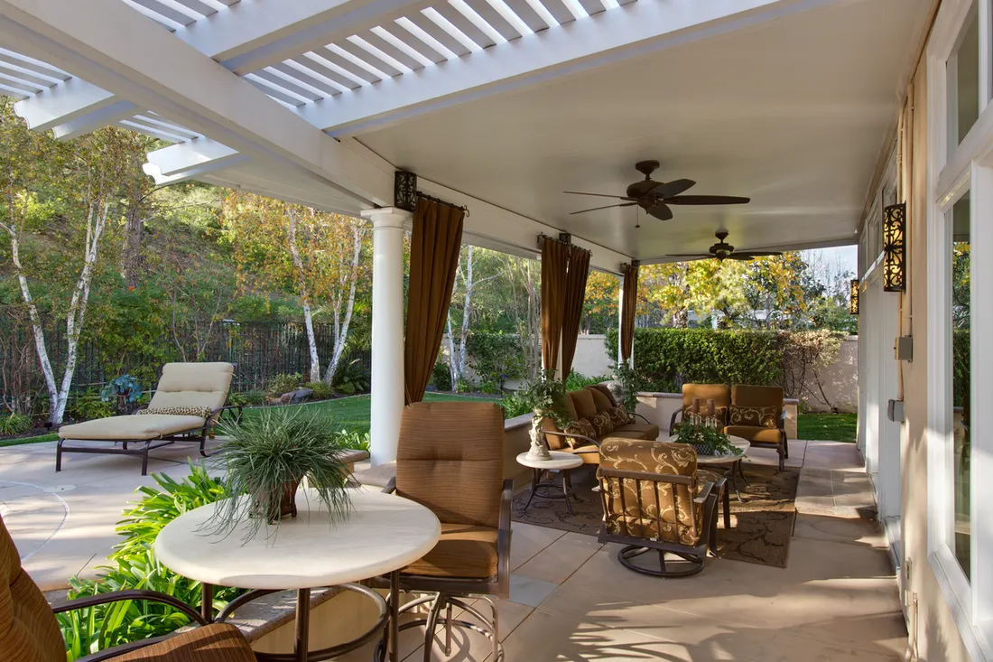 Covered patio with white columns, ceiling fan, outdoor seating, tile floor, and wooded garden surroundings