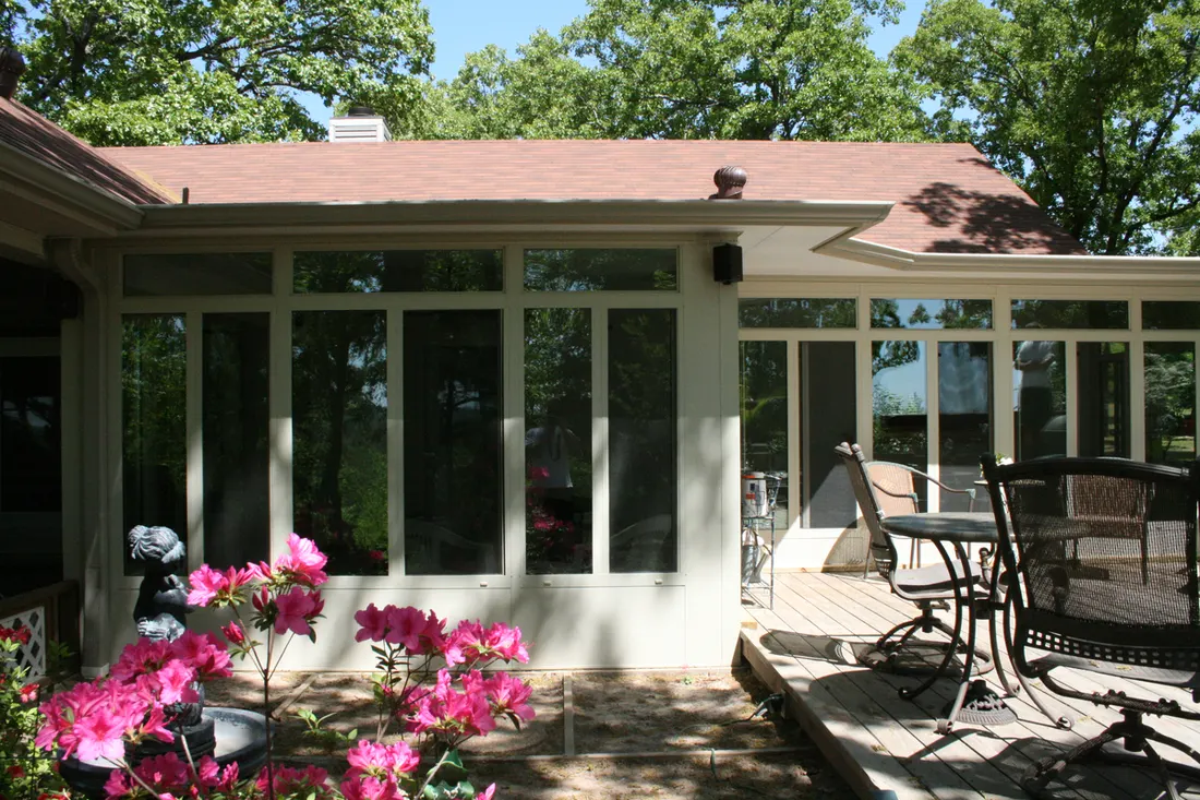 Enclosed patio sunroom with off-white framing, tall glass windows, and adjoining wooden deck patio furniture