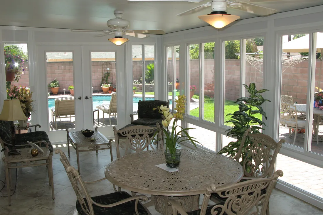 White glass patio enclosure with tile flooring and dining furniture overlooking a backyard swimming pool