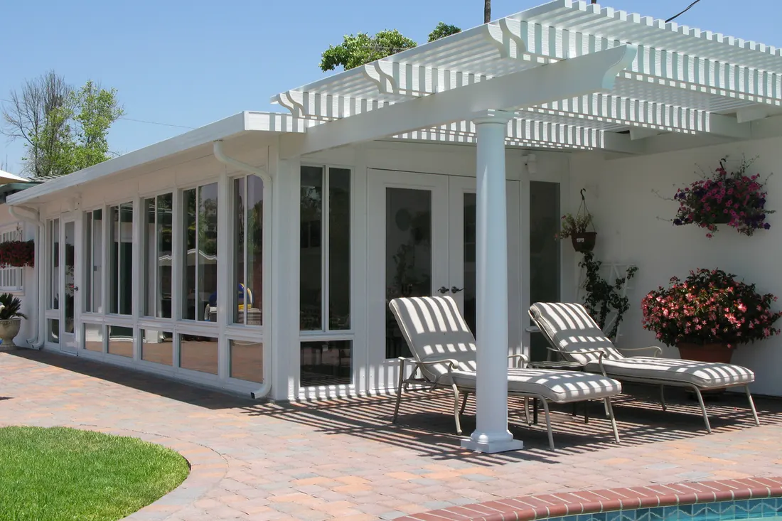 White patio enclosure with glass walls, pergola roof, lounge chairs, and flowering plants beside a brick patio