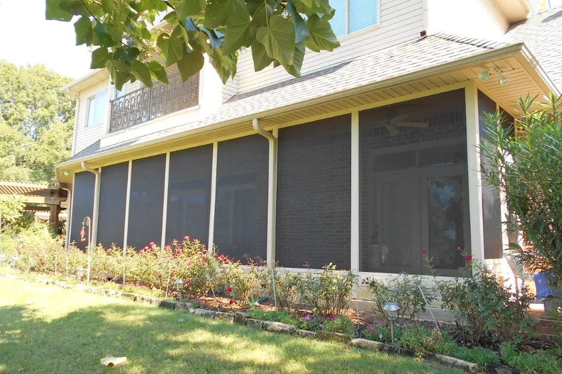Screened patio enclosure with beige framing beneath a house roofline overlooking a landscaped garden
