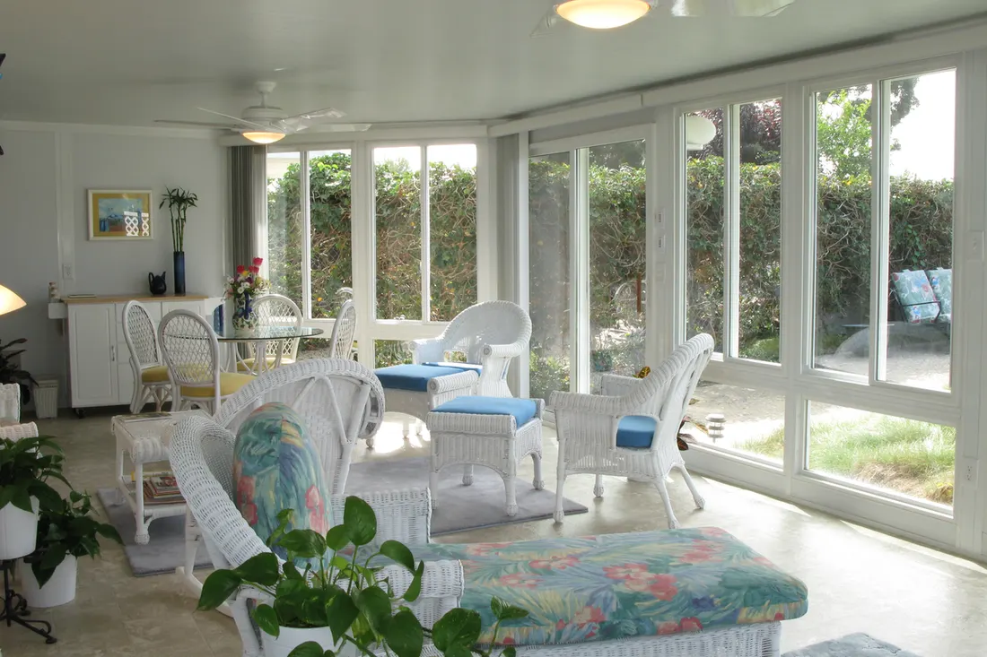 Enclosed patio room with white-framed glass walls, ceiling fans, and white wicker furniture overlooking greenery