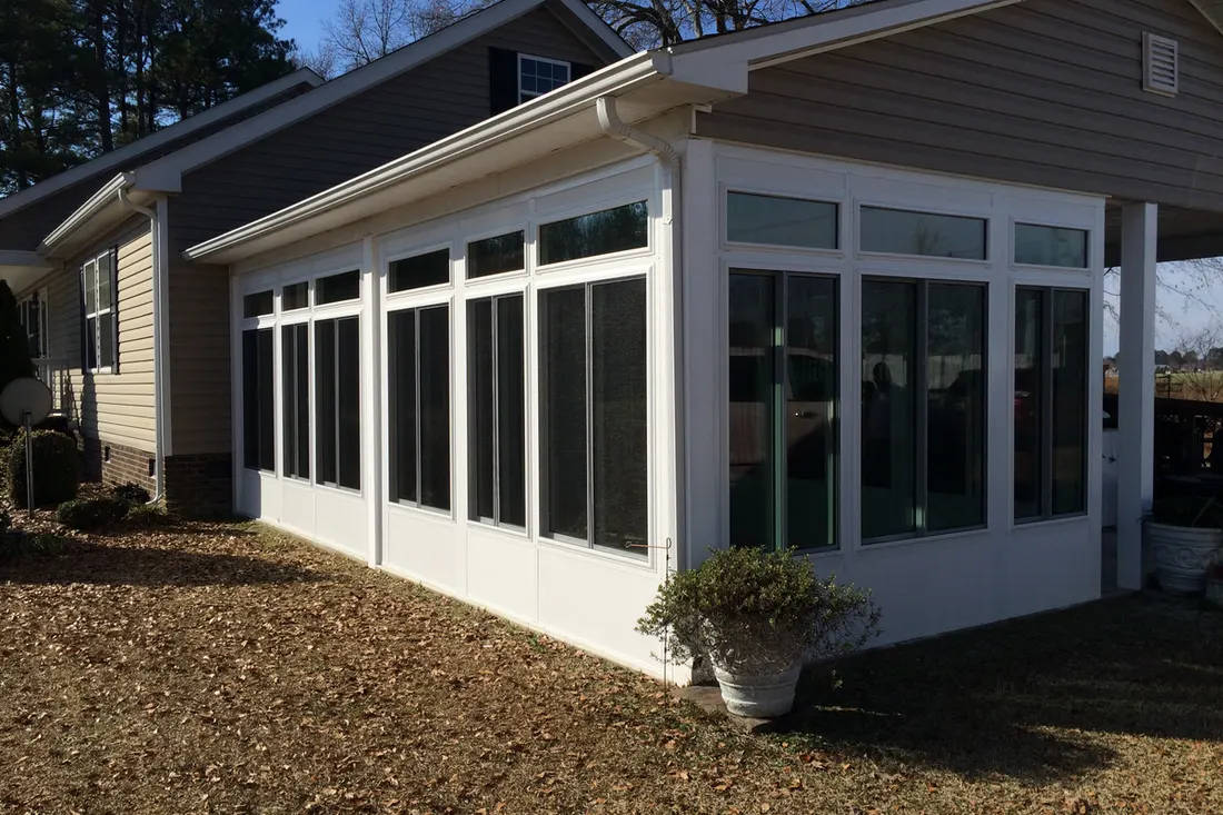 Enclosed patio with white-framed windows, gray siding, and a sloped roof beside a landscaped yard
