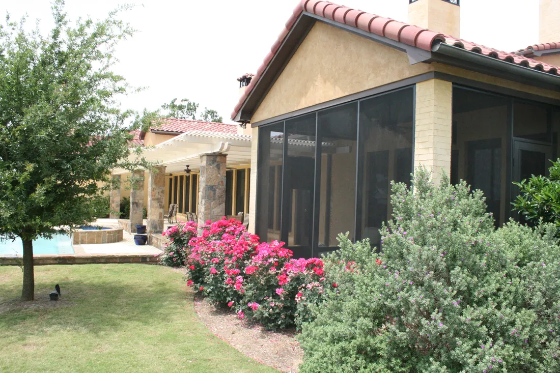 Screened patio enclosure on a stucco home with clay tile roof beside landscaped backyard bushes