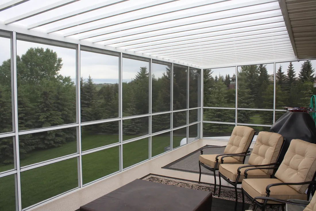 Screened patio enclosure with aluminum framing, clear panels, cushioned chairs, and wooded lawn beyond