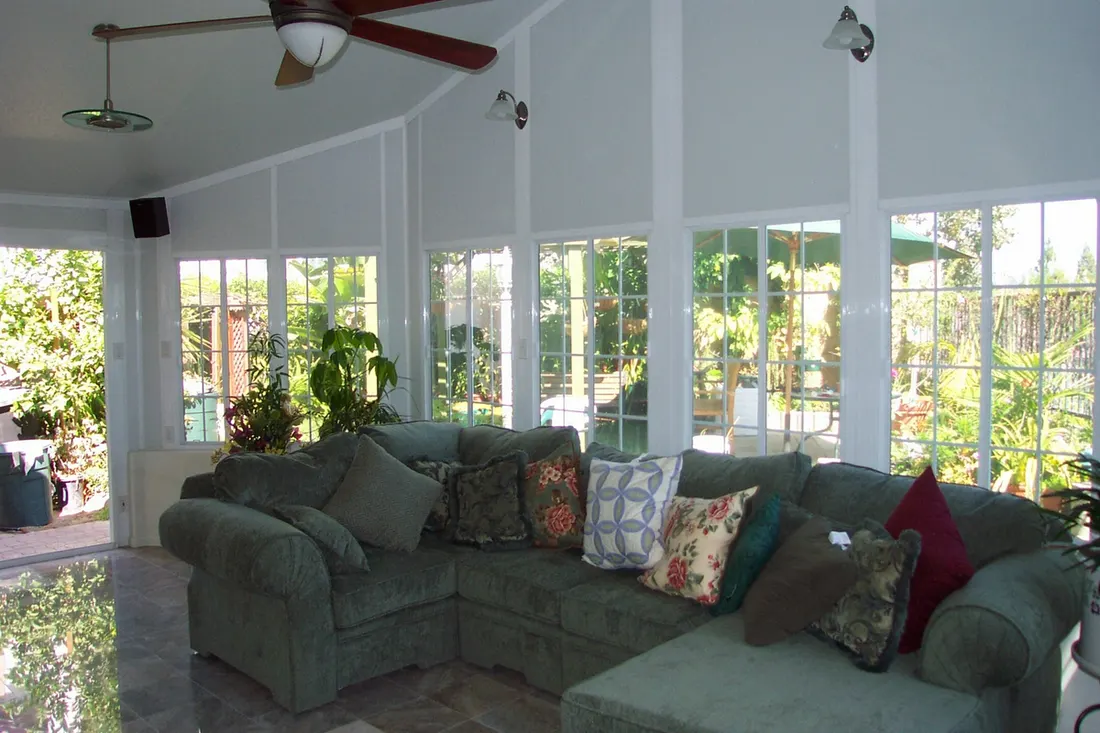 Enclosed patio sunroom with white paneled walls, grid windows, tiled floor, and green sectional sofa