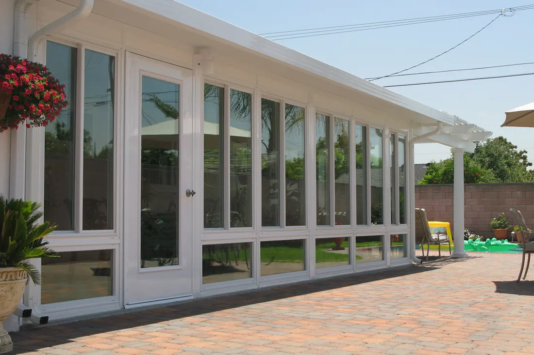 White-framed glass patio enclosure with full-height windows, brick paver floor, and flowering plants