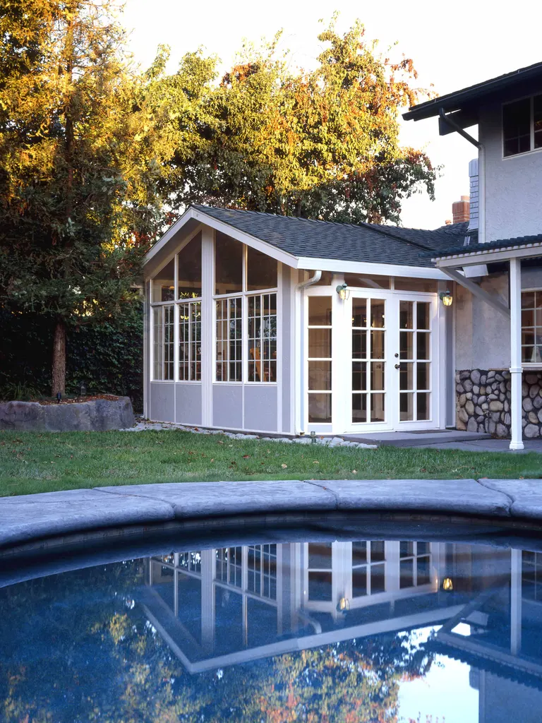 White-framed glass patio enclosure with gabled roof beside a swimming pool and landscaped yard