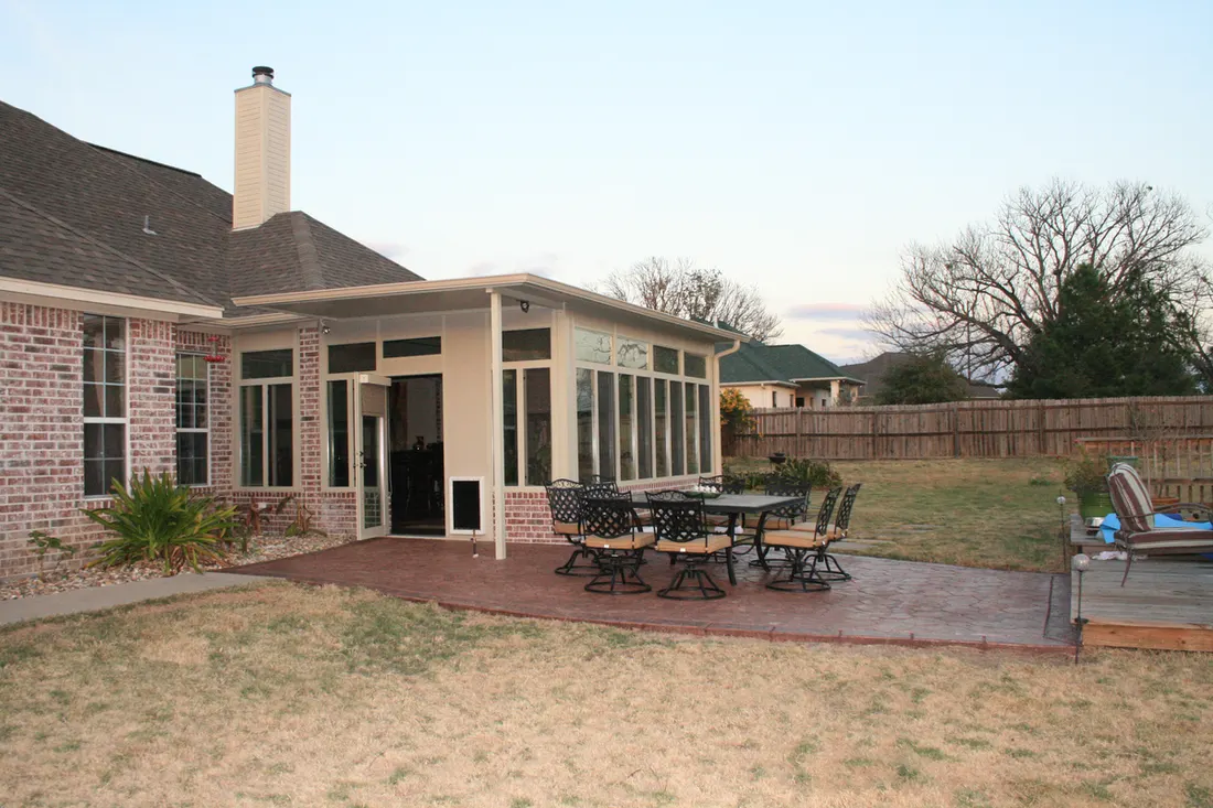 Brick patio enclosure with tall windows, beige framing, outdoor dining furniture, and a fenced yard