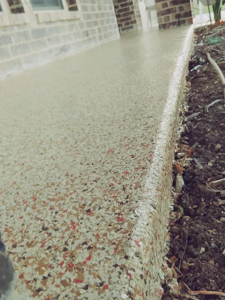 Speckled gray patio floor coating with red and tan flakes beside a brick house and garden bed