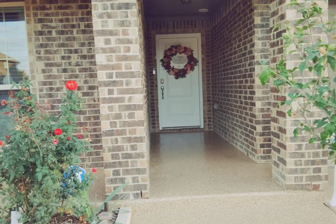 Speckled tan coated patio floor leading to a recessed entry between brick walls with flower beds