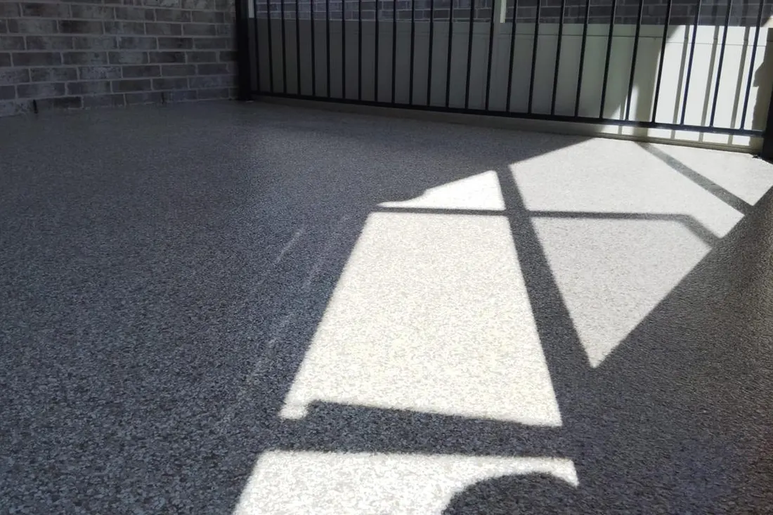 Speckled gray patio floor coating with geometric railing shadows beside a dark brick wall