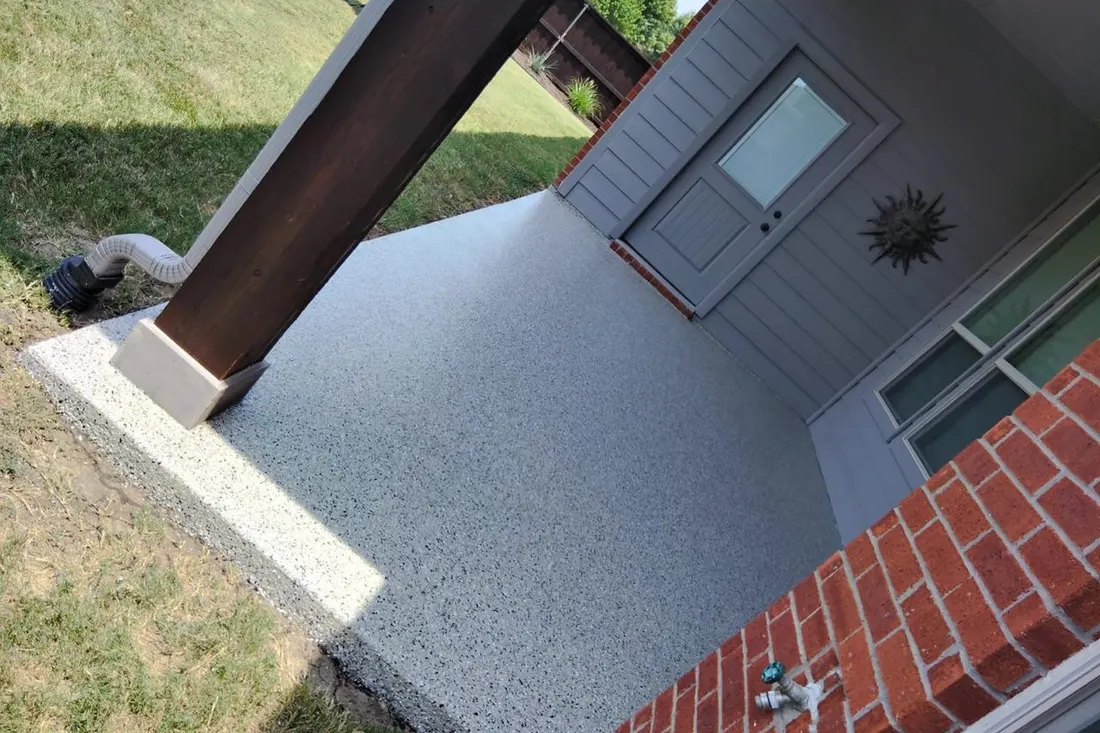 Light gray textured patio coating beside a gray-sided entry, brick wall, and wooden support post