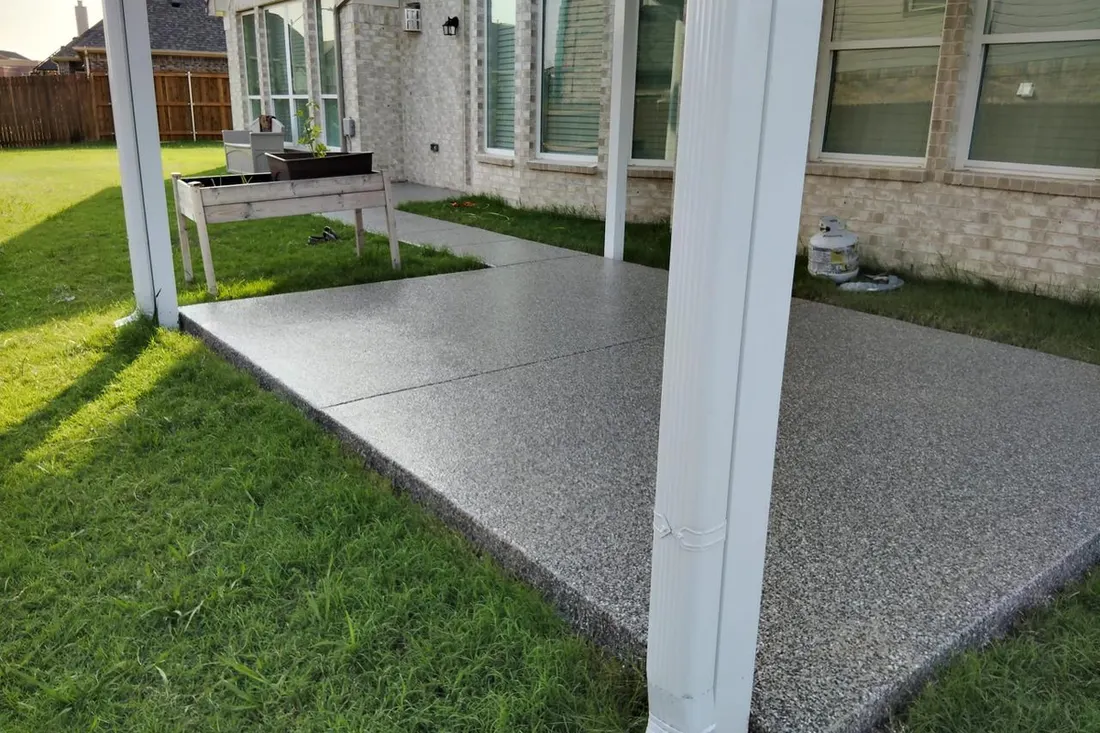 Speckled gray coated patio floor beneath a covered backyard porch beside a stone-sided house and lawn