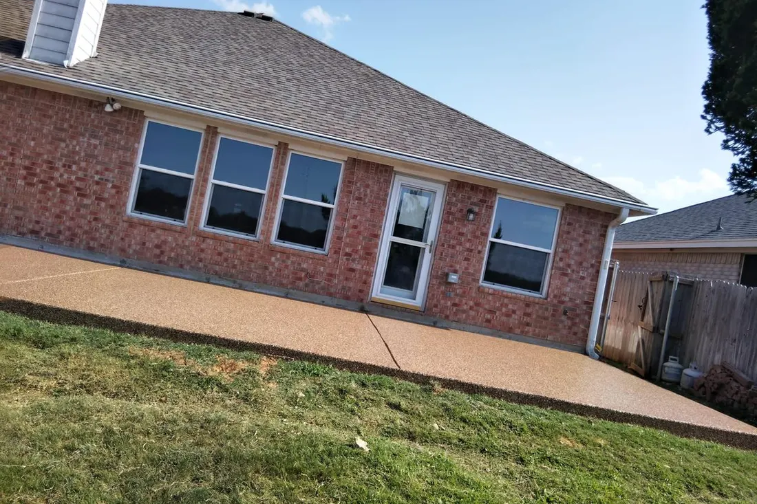 Tan textured patio coating beside a red brick home, with windows, door, lawn, and wooden fence