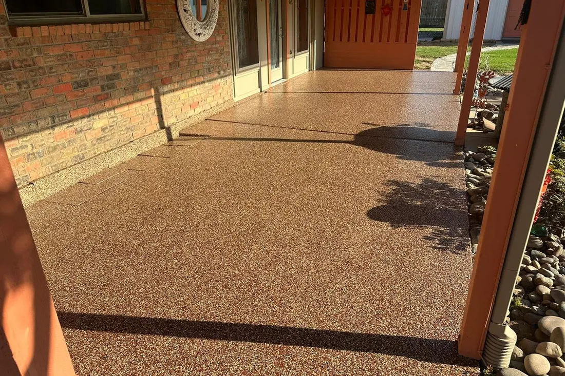 Brown textured patio floor coating beside a brick wall, glass doors, red entry door, and rock border