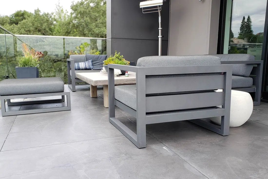 Gray coated patio floor with modern outdoor seating, glass railing, greenery, and a dark exterior wall