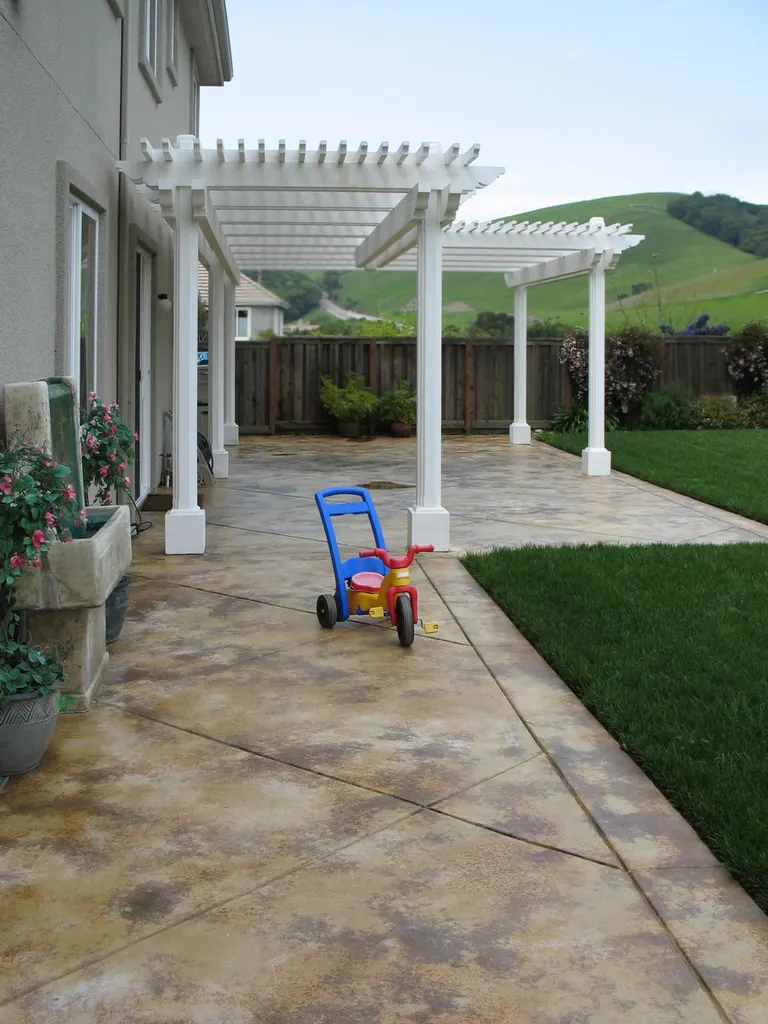 Stamped concrete patio floor in tan and gray beneath white pergolas beside a green lawn