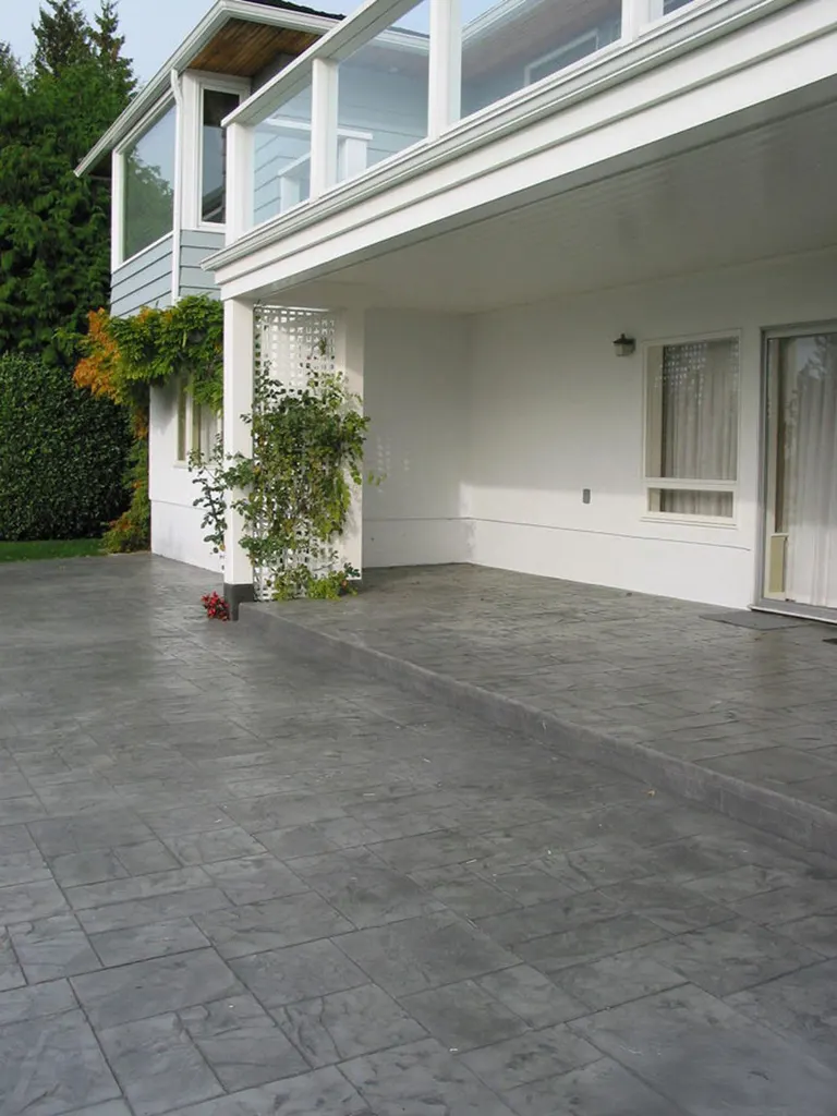 Gray stamped concrete patio floor beside a white house with windows, doors, and greenery