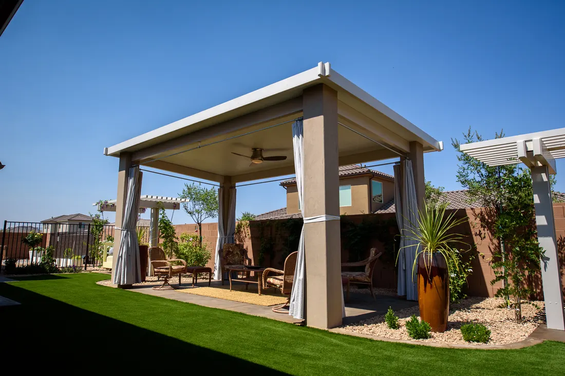 Covered beige pergola with square columns, ceiling fan, outdoor seating, and landscaped backyard