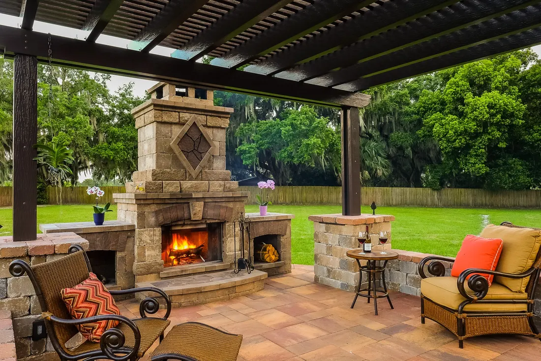 Dark wood pergola with stone fireplace, patio seating, and a green lawn surrounded by trees