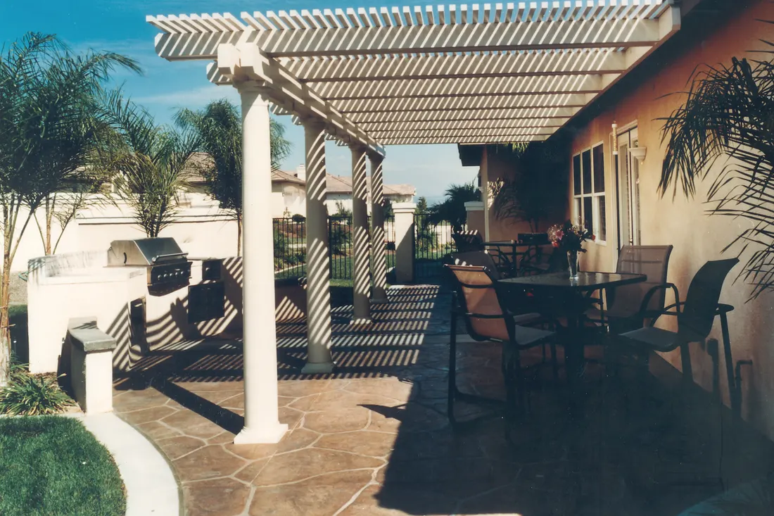 White slatted pergola over a stone patio beside a tan house, with outdoor dining and palm trees