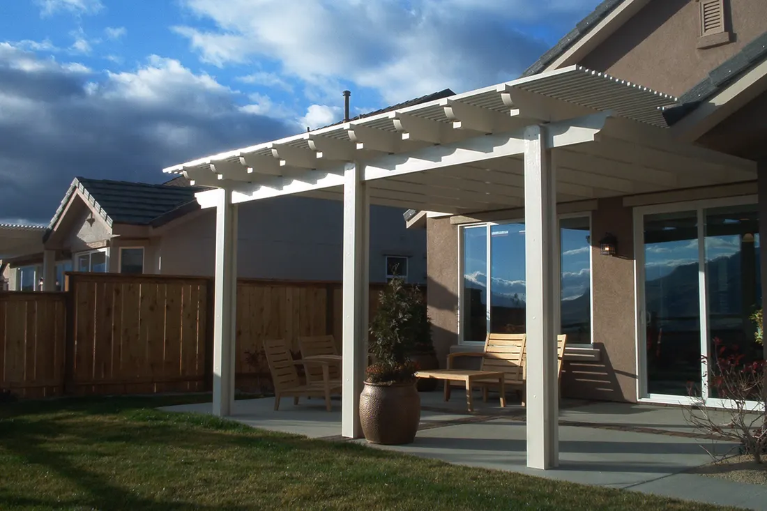 White pergola shades a patio beside a tan house, wood fence, and lawn