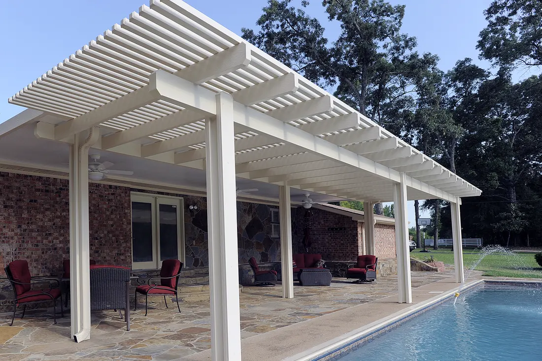 White pergola with slatted roof covers a stone patio beside a swimming pool and brick wall