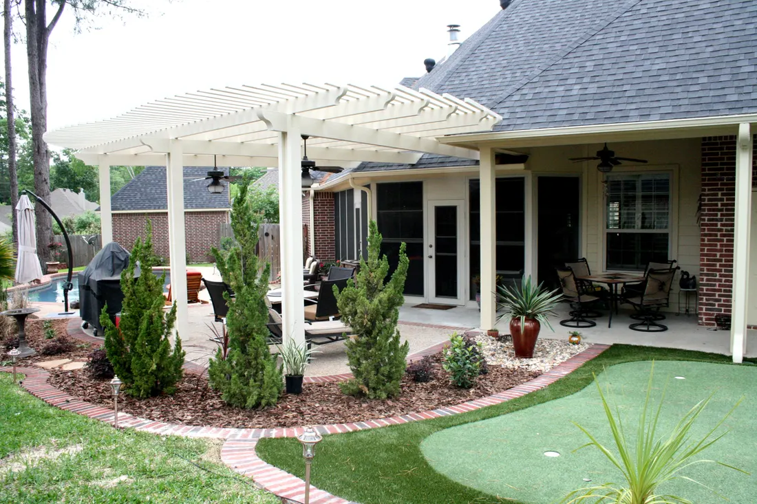 White slatted pergola beside a brick home, with patio seating, shrubs, and artificial grass