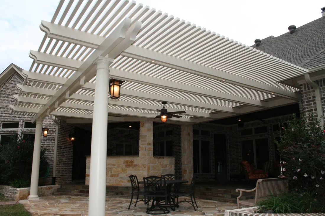 White slatted pergola with round columns over a stone patio attached to a brick house