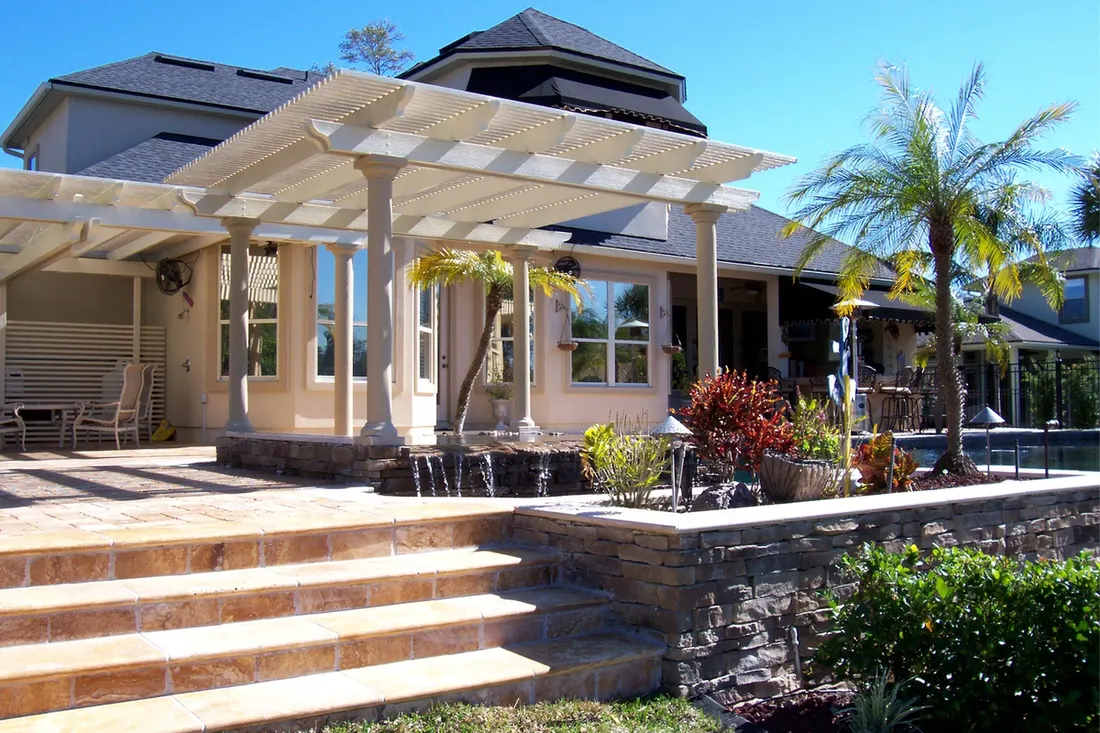 White louvered patio pergola supported by columns over a stone-paved backyard terrace with steps
