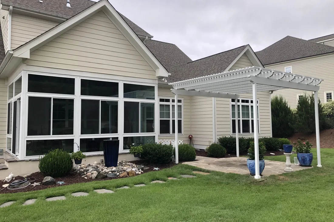 White pergola with decorative rafters beside a beige-sided house and landscaped lawn