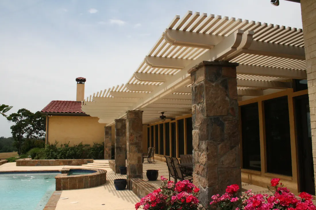Stone-columned pergola with a white slatted roof beside a swimming pool and red flowers