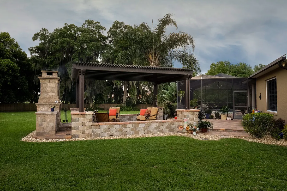Dark brown pergola over a stone retaining wall and fireplace in a green backyard