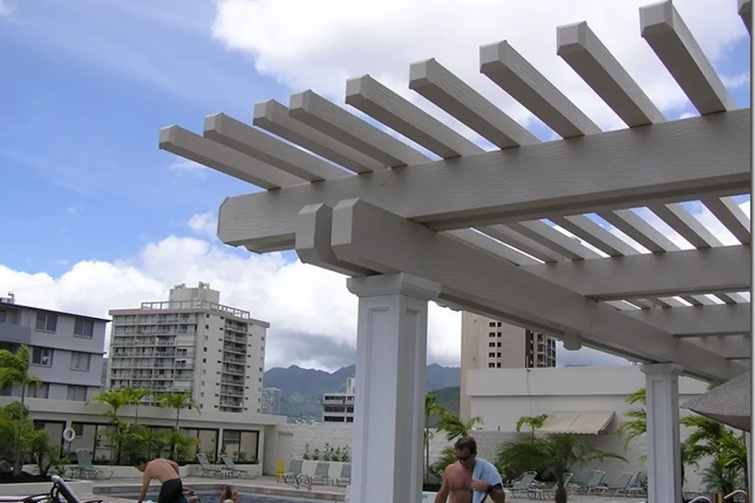 White pergola with exposed beams and slatted roof overlooking buildings and mountains under a blue sky