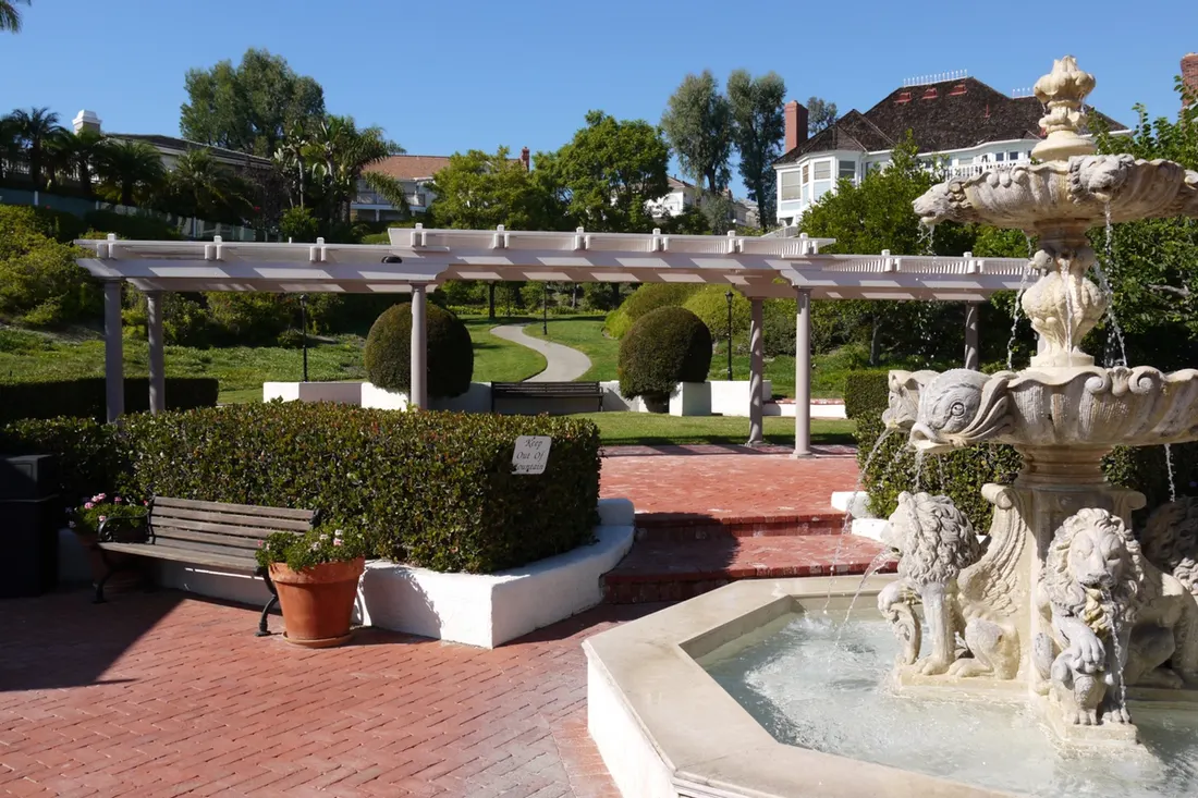White wooden pergola over a brick courtyard beside a stone fountain and manicured garden lawn