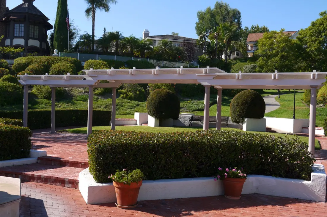 White wooden pergola with open slatted roof, manicured hedges, potted plants, and brick patio
