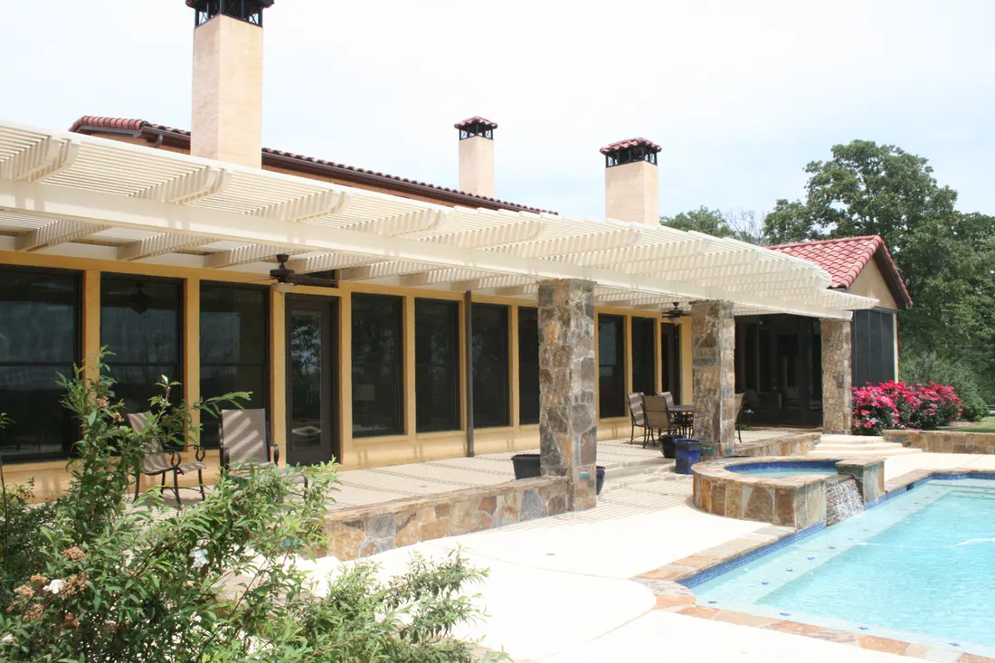 White pergola with stone columns extends over a patio beside a swimming pool