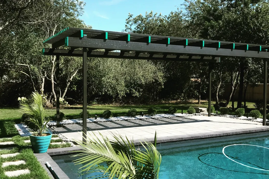 Black metal pergola over a stone paver patio beside a backyard swimming pool
