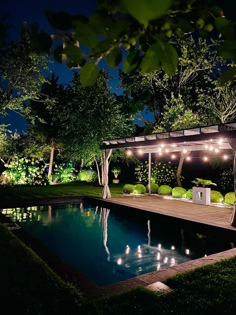 Wooden pergola with string lights beside a swimming pool, surrounded by landscaped greenery at night