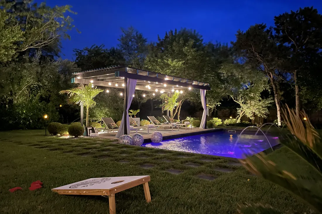 Illuminated poolside pergola with string lights, curtains, and lounge chairs at night beside a swimming pool