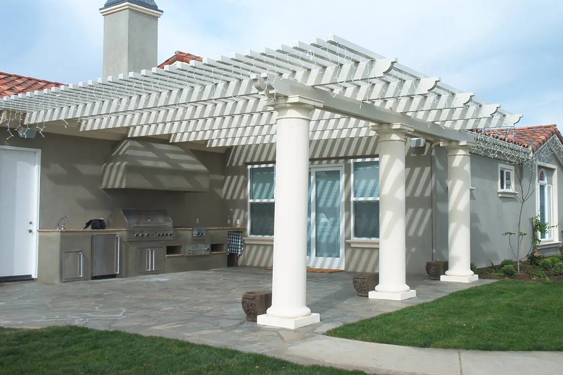 White pergola with square columns shading a stone patio beside an outdoor kitchen and stucco home