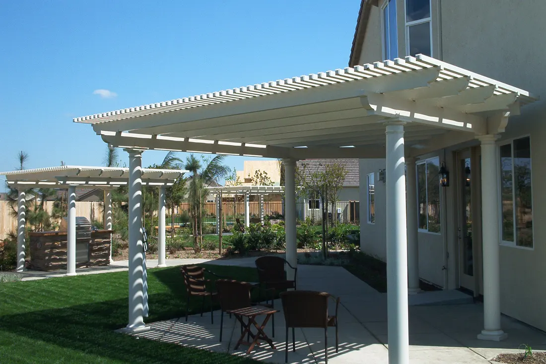 White attached pergola with round columns over a concrete patio beside a green lawn