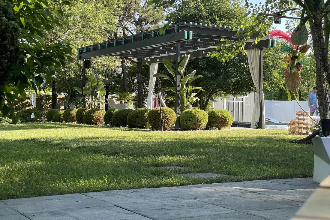 Dark metal pergola with white curtains and string lights set in a landscaped grassy backyard