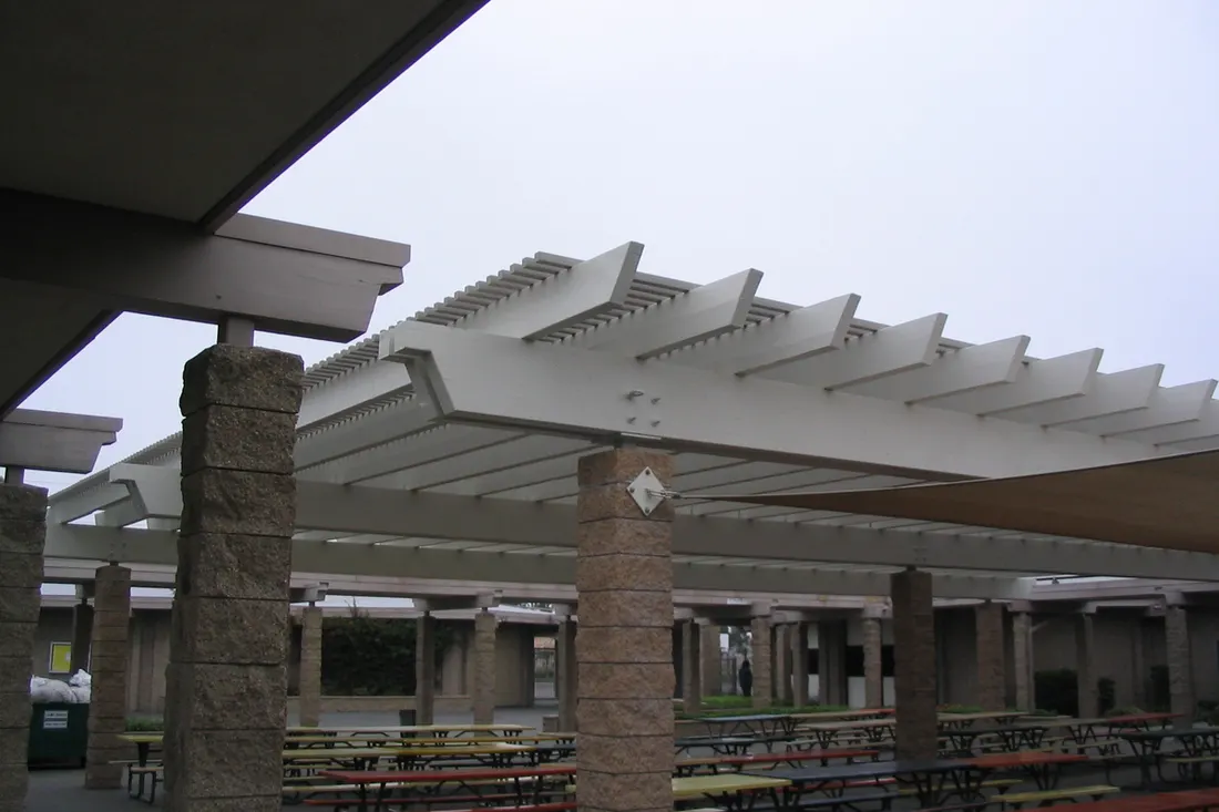 White slatted pergola supported by textured stone columns over an outdoor courtyard with picnic tables